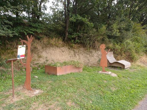 Skulpturenpark mit Metallfiguren und einer Bank aus Holz, umgeben von Gras und Bäumen.