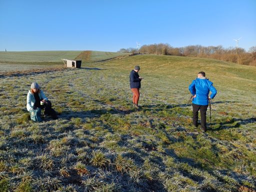 Drei Personen auf einer frostigen Wiese, eine sitzt, zwei stehen und schauen auf den Boden.