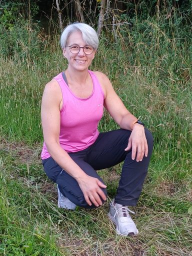 Eine Person mit grauen Haaren in einem pinken Oberteil, sitzt im Gras.