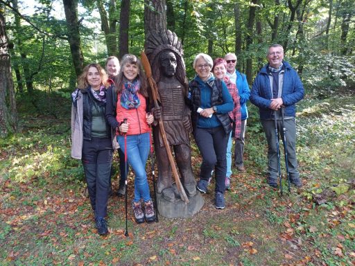Eine Gruppe von sechs Menschen steht neben einer Holzskulptur im Wald.