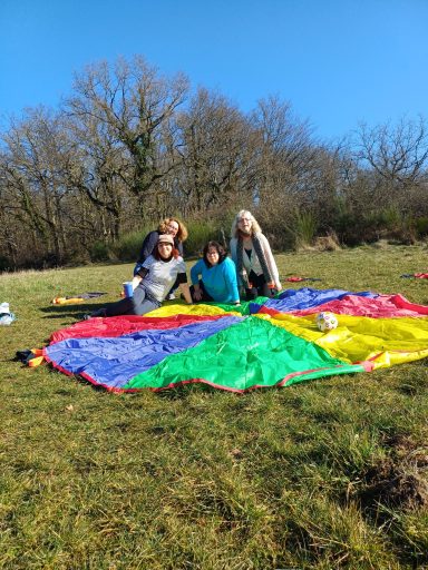 Vier Personen sitzen auf einem bunten Schwungtuch auf einer Wiese bei sonnigem Wetter.