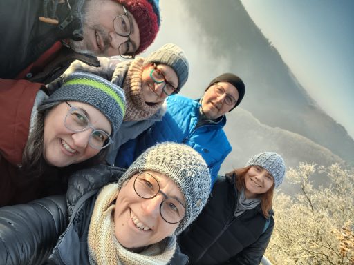 Gruppe von sechs Personen bei sonnigem Wetter im Freien, lächelnd und mit warmen Mützen.
